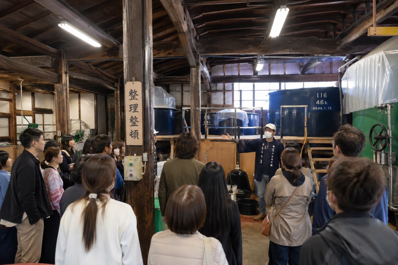 酒蔵の中で、仕込みタンクの前に立つ蔵人の説明を聞く見学ツアーの参加者